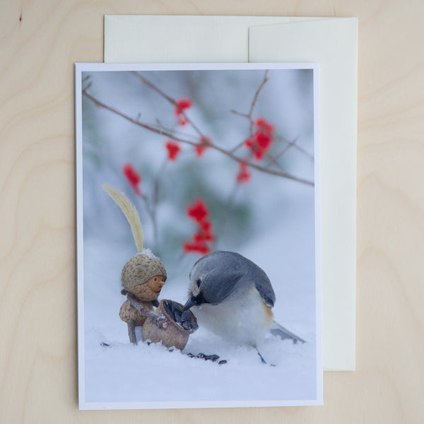 A closed greeting card with text on top of an off-white envelope on a pale wood surface. On the card is a photo of a Becorn holding a basket of seeds looking in awe at a tufted titmouse that is picking a sunflower seed out of the basket. The ground is covered in snow, and there are a few bright red berries in the background.