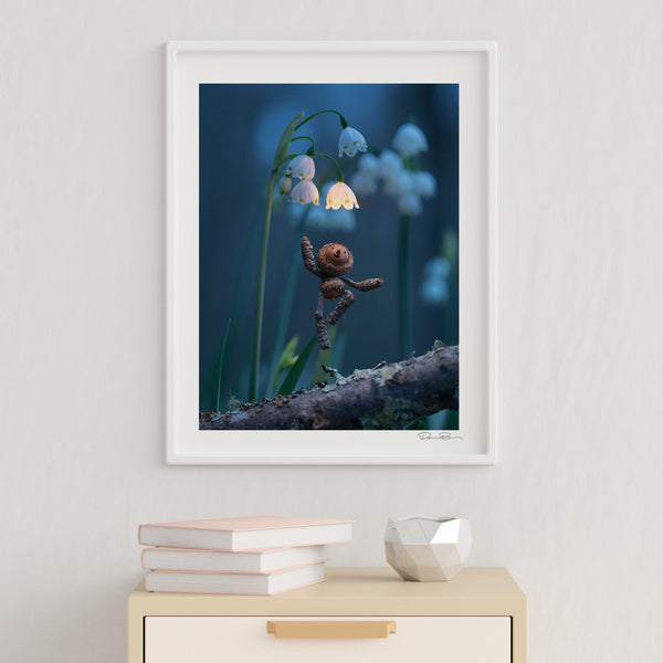 The photo of a Becorn underneath snowbells in a thin, white frame hanging on an off-white wall. Underneath it is a small table with an accent bowl and a small stack of books. In the lower right corner of the print’s white border is David Bird’s signature.