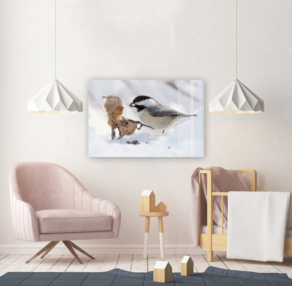 The photo of a Becorn and chickadee in the snow as a metal print, mounted without a frame on an off-white wall. The metal print faintly reflects an unseen plain wall. Underneath it is a wooden chair and crib, and it is bracketed by white hanging lamps.