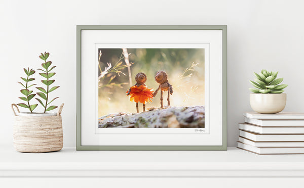 The photo of two Becorns holding hands in a thin, sage green frame on a white shelf against a white wall. On either side of it are potted plants and a small stack of books. In the lower right corner of the print’s white border is David Bird’s signature.