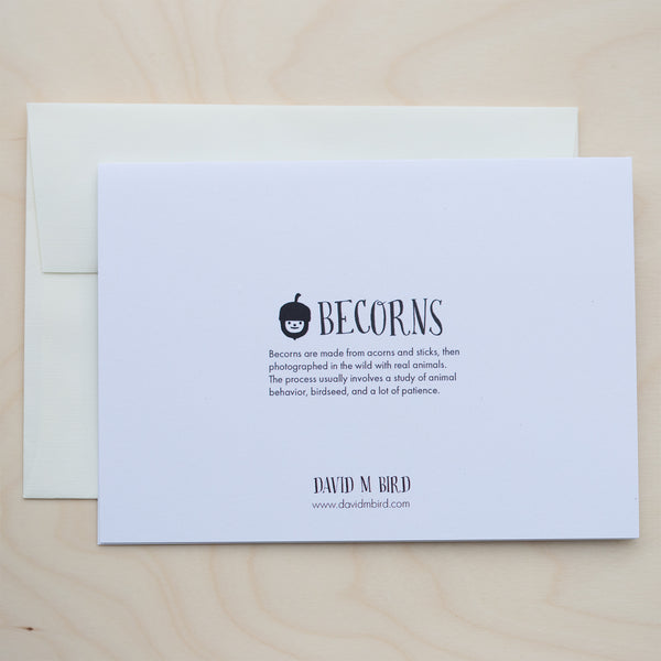 A closed, white greeting card with text on top of an off-white envelope on a pale wood surface. Logo: A black and white acorn with a smiley face. Text: Becorns. Becorns are made with acorns and sticks, then photographed in the wild with real animals. The process usually involves a study of animal behavior, birdseed, and a lot of patience. David M Bird. www.davidmbird.com