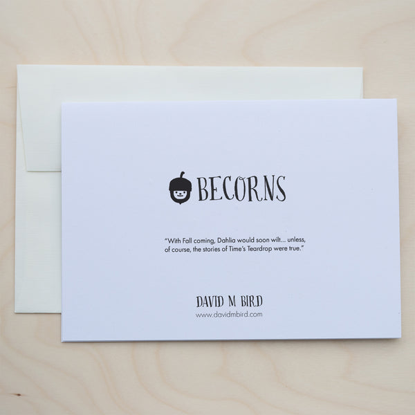 A closed, white greeting card with text on top of an off-white envelope on a pale wood surface. Logo: A black and white acorn with a smiley face. Text: Becorns. “With Fall coming, Dahlia would soon wilt… unless, of course, the stories of Time’s Teardrop were true.” David M Bird. www.davidmbird.com