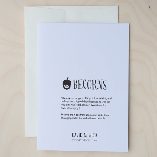 A closed, white greeting card with text on top of an off-white envelope on a pale wood surface. Logo: A black and white acorn with a smiley face. Text: Becorns. “There was a magic to this spot. Joonie felt it, and perhaps the chippy did too because he was out way past his usual bedtime.” (Watch for owls, little chippy!) Becorns are made with acorns and sticks, then photographed in the wild with real animals. David M Bird. www.davidmbird.com